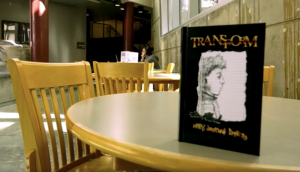 A book titled "TRANSFORM" with a black cover and an illustration of a person's profile is displayed upright on a round table in at a college of the canyons study area. The background features wooden chairs, tables, large windows, and a person using a laptop.